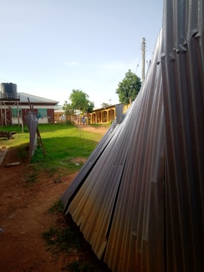 Corrugated metal sheets are stacked beside a path leading to a row of buildings with a water tank on stilts nearby. There are a few small trees and grass lawns along the path, and power lines are visible above. The scene is lit by natural daylight under a clear sky.
