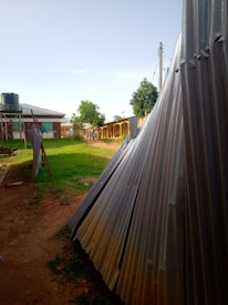 Corrugated metal sheets are stacked beside a path leading to a row of buildings with a water tank on stilts nearby. There are a few small trees and grass lawns along the path, and power lines are visible above. The scene is lit by natural daylight under a clear sky.