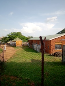 A chain-link fence encloses an area with a brick building to the right, labeled as a mental health facility. In the center, there is a small, bright yellow kiosk or shed with a green roof. The area is grassy with a dirt path leading towards the kiosk, and several trees provide greenery in the background. The sky is clear with a few scattered clouds, suggesting pleasant weather.