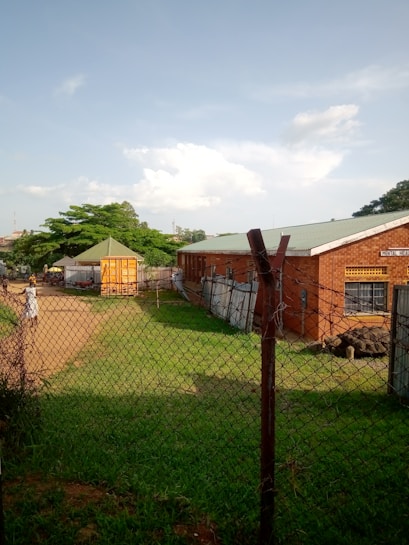 A chain-link fence encloses an area with a brick building to the right, labeled as a mental health facility. In the center, there is a small, bright yellow kiosk or shed with a green roof. The area is grassy with a dirt path leading towards the kiosk, and several trees provide greenery in the background. The sky is clear with a few scattered clouds, suggesting pleasant weather.