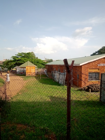 A chain-link fence encloses an area with a brick building to the right, labeled as a mental health facility. In the center, there is a small, bright yellow kiosk or shed with a green roof. The area is grassy with a dirt path leading towards the kiosk, and several trees provide greenery in the background. The sky is clear with a few scattered clouds, suggesting pleasant weather.