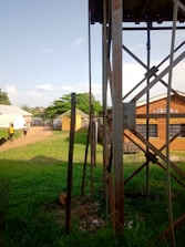 A grassy area with a tall metal structure in the foreground, possibly a water tower, with a ladder attached. There are people walking on a dirt path that runs through the scene. The path leads to several buildings with varied roofing, one of which is marked as a mental health unit. Trees and other vegetation are visible in the background under a partly cloudy sky.
