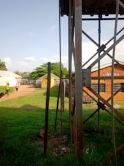 A grassy area with a tall metal structure in the foreground, possibly a water tower, with a ladder attached. There are people walking on a dirt path that runs through the scene. The path leads to several buildings with varied roofing, one of which is marked as a mental health unit. Trees and other vegetation are visible in the background under a partly cloudy sky.