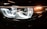 Close-up view of a car's headlight, featuring intricate LED design and reflective surfaces. The light emits a bright, white glow, accented by amber lighting on the side.