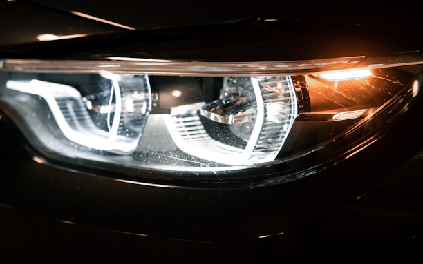 Close-up view of a car's headlight, featuring intricate LED design and reflective surfaces. The light emits a bright, white glow, accented by amber lighting on the side.