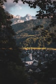 A picturesque valley town nestled between towering evergreen forests and dramatic mountain peaks bathed in soft light. A prominent church with a tall steeple stands out among the clustered houses. Leaves from nearby trees frame the scene, adding a sense of depth and serenity.