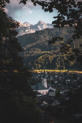 A picturesque valley town nestled between towering evergreen forests and dramatic mountain peaks bathed in soft light. A prominent church with a tall steeple stands out among the clustered houses. Leaves from nearby trees frame the scene, adding a sense of depth and serenity.