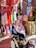 An elderly woman dressed in traditional clothing with vibrant floral patterns sits beside a small market stall. Surrounding her are colorful scarves and fabrics hanging on display. There are also plants and possibly goods for sale beside her.