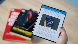 A person is holding a folding smartphone with the screen unfolded, displaying a webpage with an image of a similar foldable phone. In the background are three stacked books with colorful covers.