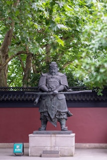 A large stone statue of a warrior stands on a pedestal. The warrior is clad in traditional armor and holds a large sword. There are lush green trees above and a tiled wall in the background, along with a small sign at the base of the statue.