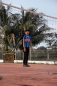 A young boy stands on a clay-colored sports court, wearing a blue sports jersey and black pants. Behind him, tall palm trees and a metal fence form the backdrop. The mood appears calm and serene under the bright daylight.