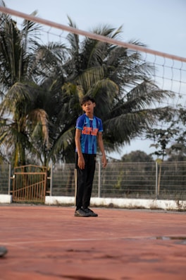 A young boy stands on a clay-colored sports court, wearing a blue sports jersey and black pants. Behind him, tall palm trees and a metal fence form the backdrop. The mood appears calm and serene under the bright daylight.