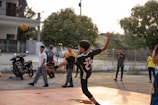A young person wearing a sports jersey is performing a high kick with a woven ball in mid-air on a court. Several other youths are standing nearby, watching the action. Motorbikes and trees are in the background, with the setting sun casting warm light.