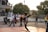 A young person wearing a sports jersey is performing a high kick with a woven ball in mid-air on a court. Several other youths are standing nearby, watching the action. Motorbikes and trees are in the background, with the setting sun casting warm light.
