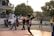 A young person wearing a sports jersey is performing a high kick with a woven ball in mid-air on a court. Several other youths are standing nearby, watching the action. Motorbikes and trees are in the background, with the setting sun casting warm light.