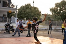 A young person wearing a sports jersey is performing a high kick with a woven ball in mid-air on a court. Several other youths are standing nearby, watching the action. Motorbikes and trees are in the background, with the setting sun casting warm light.