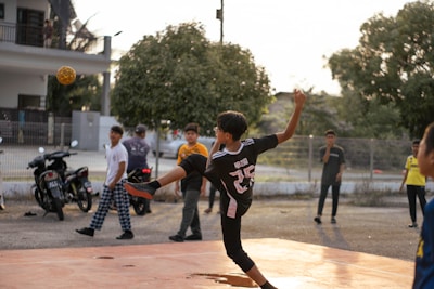 A young person wearing a sports jersey is performing a high kick with a woven ball in mid-air on a court. Several other youths are standing nearby, watching the action. Motorbikes and trees are in the background, with the setting sun casting warm light.