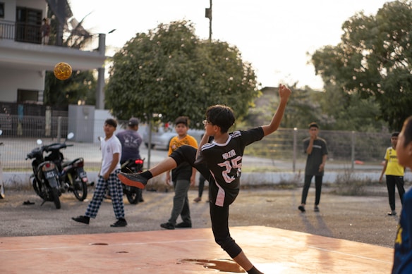 A young person wearing a sports jersey is performing a high kick with a woven ball in mid-air on a court. Several other youths are standing nearby, watching the action. Motorbikes and trees are in the background, with the setting sun casting warm light.