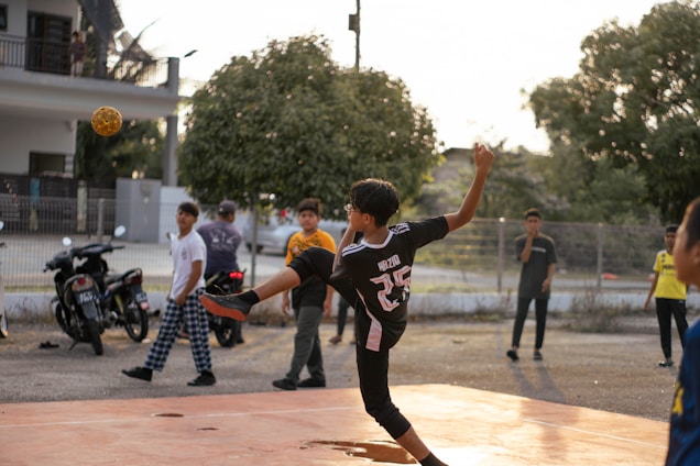 A young person wearing a sports jersey is performing a high kick with a woven ball in mid-air on a court. Several other youths are standing nearby, watching the action. Motorbikes and trees are in the background, with the setting sun casting warm light.