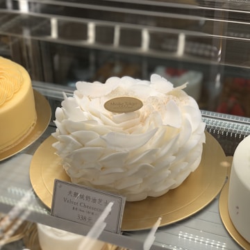 A sophisticated white cake designed with intricate petal-like decorations, displayed on a golden cake board. Positioned inside a glass display case, there are other cakes in the background, one of which is a light yellow, rosette-patterned cake. An elegant label on the white cake reads 'Mvuke Tokyo,' and a placard in front of it details that it is a Velvet Cheesecake priced at 338 yuan.