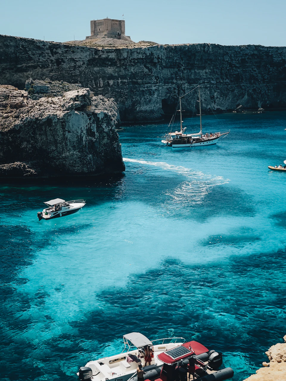 The image depicts a serene coastal scene with several boats floating on the water, including a white boat and a red boat. The boats are scattered across the deep blue waters, creating a sense of depth and perspective. In the background, there is a cliff made of gray rocks that adds to the rugged beauty of the landscape. A few people can be seen standing near these rocks, possibly enjoying the view or preparing for their next adventure on the water. The sky above them is clear and blue, suggesting a perfect day for boating activities.