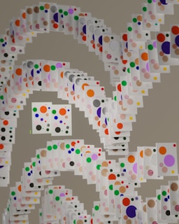 A creative arrangement of white cards, each adorned with colorful dots in varying sizes. These cards are suspended in a spiral formation creating a dynamic and layered visual effect. The dots on the cards are red, blue, green, orange, purple, pink, yellow, and black, contrasting against the white background of the cards.