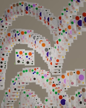 A creative arrangement of white cards, each adorned with colorful dots in varying sizes. These cards are suspended in a spiral formation creating a dynamic and layered visual effect. The dots on the cards are red, blue, green, orange, purple, pink, yellow, and black, contrasting against the white background of the cards.