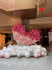 A large heart shape made out of various sizes of pink and white balloons is positioned indoors. The balloons are stacked together, creating a prominent 3D structure. The heart is surrounded by additional white and pink balloons on the floor. The setting appears to be in a commercial indoor space, indicated by the visible store signage and the modern architectural elements around the display.