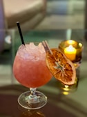 A pink cocktail in a rounded glass with crushed ice. It features a dried orange slice and a small clip as a garnish. A black straw is inserted in the drink. In the background, a lit candle inside a holder adds a warm glow, contributing to an elegant and cozy atmosphere.