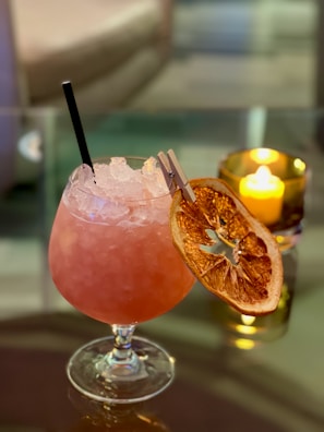 A pink cocktail in a rounded glass with crushed ice. It features a dried orange slice and a small clip as a garnish. A black straw is inserted in the drink. In the background, a lit candle inside a holder adds a warm glow, contributing to an elegant and cozy atmosphere.