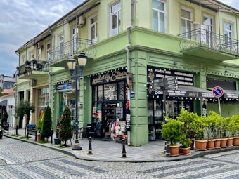 A corner building with a faded green exterior featuring wrought iron balconies, situated at a cobblestone street intersection. There are shopfronts on the ground floor with signs referencing Georgian wine and cheese. Potted plants line the entrance alongside a few street lamps. The sky above is overcast.