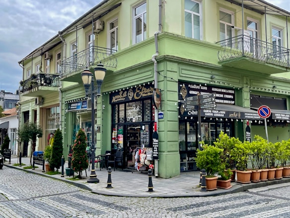 A corner building with a faded green exterior featuring wrought iron balconies, situated at a cobblestone street intersection. There are shopfronts on the ground floor with signs referencing Georgian wine and cheese. Potted plants line the entrance alongside a few street lamps. The sky above is overcast.