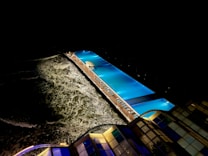 An illuminated swimming pool adjacent to a rough ocean creates a striking contrast against the dark night sky. The scene captures the gentle waves and foamy surf of the sea, with the glow from the pool lights reflecting off the water's surface. Architectural structures with glass panels are also visible, complemented by the serene ambiance of the pool area unknown to any human activity.