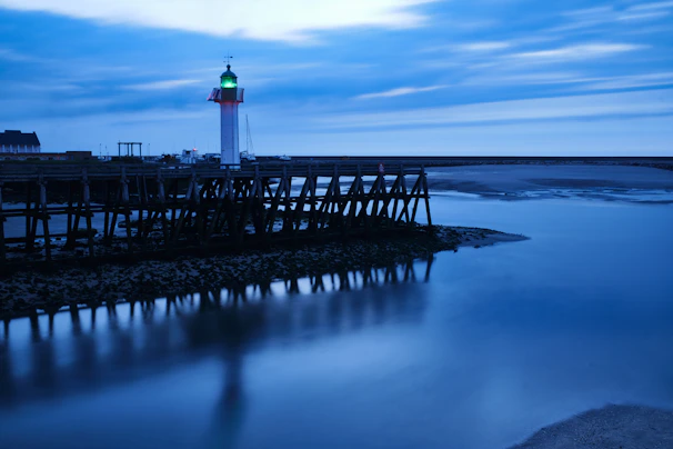 A serene navy blue lighthouse silhouette with golden beams shining across a calm sea at dusk.