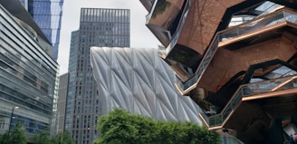 A modern urban scene featuring uniquely designed buildings with a mix of glass, metal, and copper elements. The architecture includes a structure with diagonal metal panels, a mirrored copper-colored building with intricate angles, and a tower with a grid of windows. Green trees are visible in the foreground.