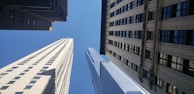 Several tall skyscrapers converge towards a bright blue sky in a cityscape with buildings of varying architectural styles.