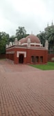 A small, red-brick mosque with multiple domes and arched doorways set in a garden area with surrounding tall palm trees and other greenery. The red structure contrasts with the green grass, and the sky is overcast, suggesting an overcast day.