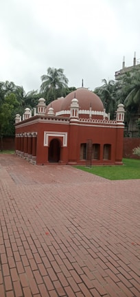 A small, red-brick mosque with multiple domes and arched doorways set in a garden area with surrounding tall palm trees and other greenery. The red structure contrasts with the green grass, and the sky is overcast, suggesting an overcast day.