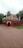 A small, red-brick mosque with multiple domes and arched doorways set in a garden area with surrounding tall palm trees and other greenery. The red structure contrasts with the green grass, and the sky is overcast, suggesting an overcast day.