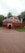 A small, red-brick mosque with multiple domes and arched doorways set in a garden area with surrounding tall palm trees and other greenery. The red structure contrasts with the green grass, and the sky is overcast, suggesting an overcast day.