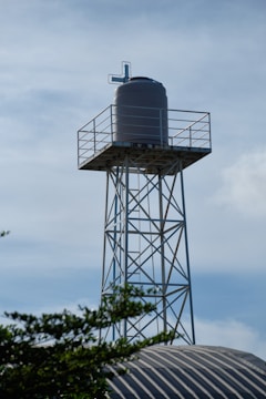 A large cylindrical water tank is mounted on a tall metal tower. The tower is constructed from steel beams forming a lattice structure. The top of the tank features a pipe or vent system. In the foreground, some foliage and the top of a corrugated metal roof are visible against a backdrop of a lightly clouded sky.