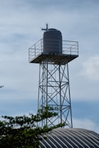 A large cylindrical water tank is mounted on a tall metal tower. The tower is constructed from steel beams forming a lattice structure. The top of the tank features a pipe or vent system. In the foreground, some foliage and the top of a corrugated metal roof are visible against a backdrop of a lightly clouded sky.