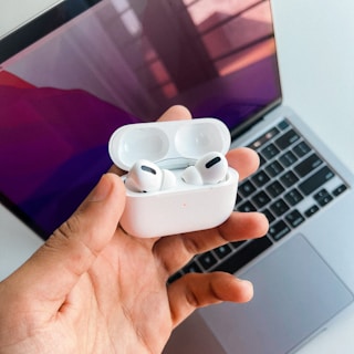 A hand holding a white pair of wireless earbuds with an open charging case, positioned in front of a laptop. The laptop screen displays a gradient of purple and pink colors, with a reflection visible on the screen.