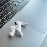 A pair of white wireless earphones resting on the silver trackpad of a laptop, alongside the keyboard. The earphones have a sleek and modern design, with black accent details. The laptop's keyboard is visible, featuring black keys with white letters.
