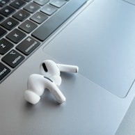 A pair of white wireless earphones resting on the silver trackpad of a laptop, alongside the keyboard. The earphones have a sleek and modern design, with black accent details. The laptop's keyboard is visible, featuring black keys with white letters.