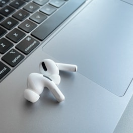 A pair of white wireless earphones resting on the silver trackpad of a laptop, alongside the keyboard. The earphones have a sleek and modern design, with black accent details. The laptop's keyboard is visible, featuring black keys with white letters.