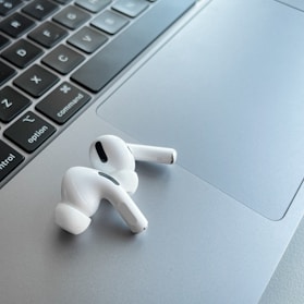 A pair of white wireless earphones resting on the silver trackpad of a laptop, alongside the keyboard. The earphones have a sleek and modern design, with black accent details. The laptop's keyboard is visible, featuring black keys with white letters.