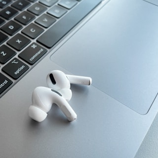 A pair of white wireless earphones resting on the silver trackpad of a laptop, alongside the keyboard. The earphones have a sleek and modern design, with black accent details. The laptop's keyboard is visible, featuring black keys with white letters.