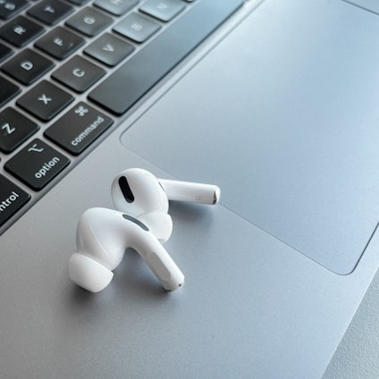A pair of white wireless earphones resting on the silver trackpad of a laptop, alongside the keyboard. The earphones have a sleek and modern design, with black accent details. The laptop's keyboard is visible, featuring black keys with white letters.
