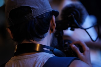 A person wearing a cap is holding a camera with a microphone attached, focusing intently on something in front of them. The scene appears to be low-lit, suggesting a nighttime or indoor setting.